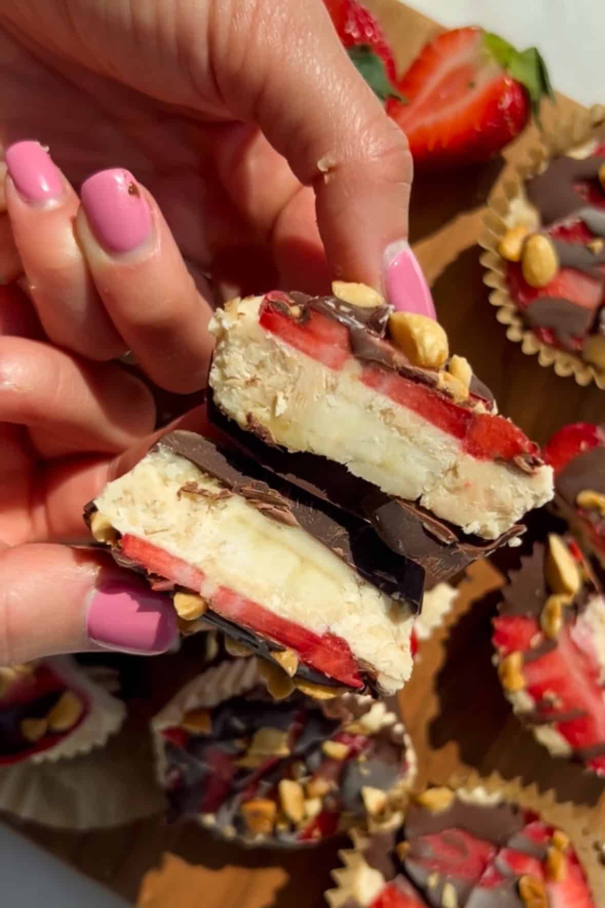 A person holding two halves of a chocolate-covered treat with visible layers of banana, strawberries, nuts, and chocolate. Other similar treats are on a tray below.