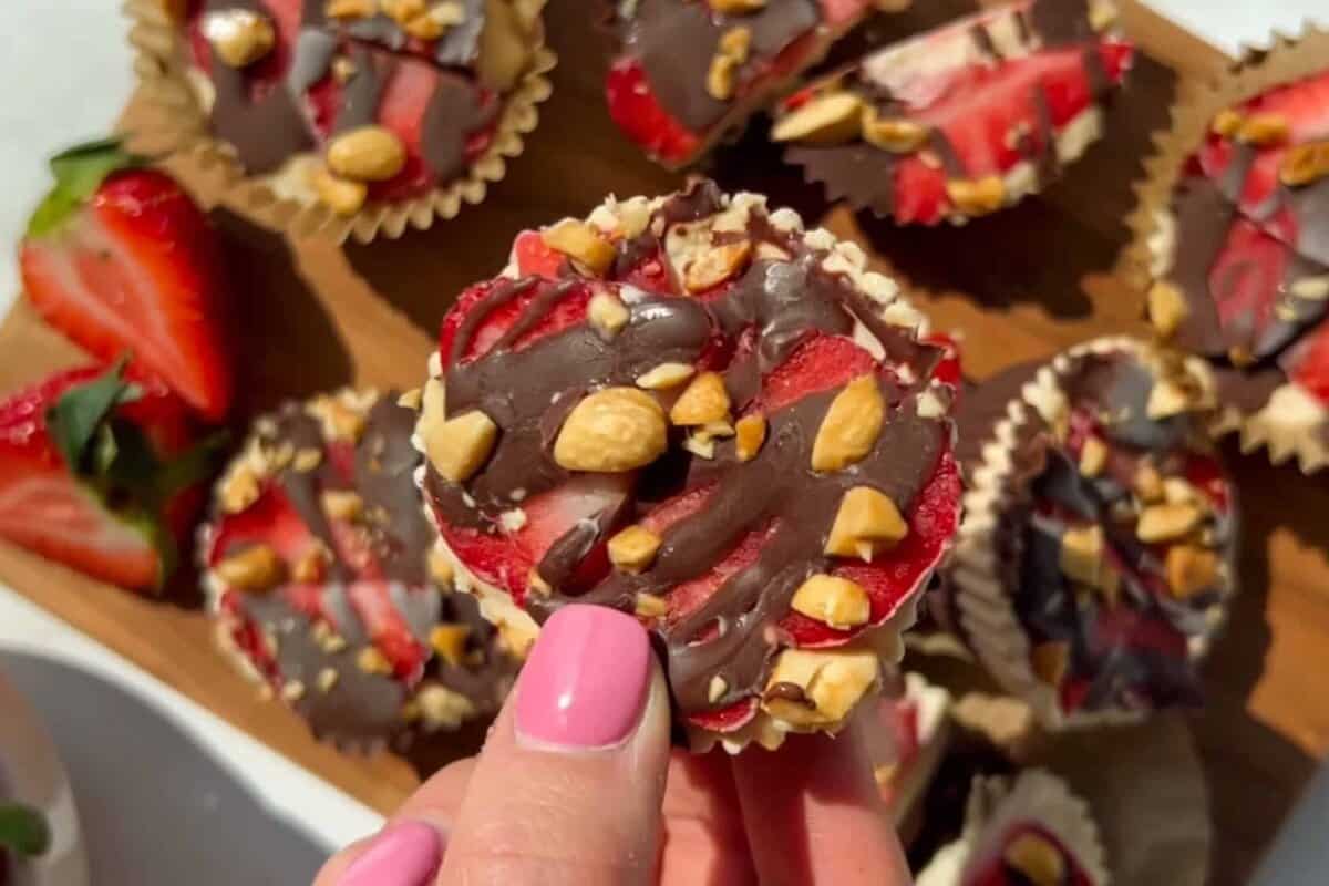 A hand with pink nail polish holds a chocolate-covered treat topped with chopped nuts and strawberry slices, with more treats and strawberries visible on a wooden tray.