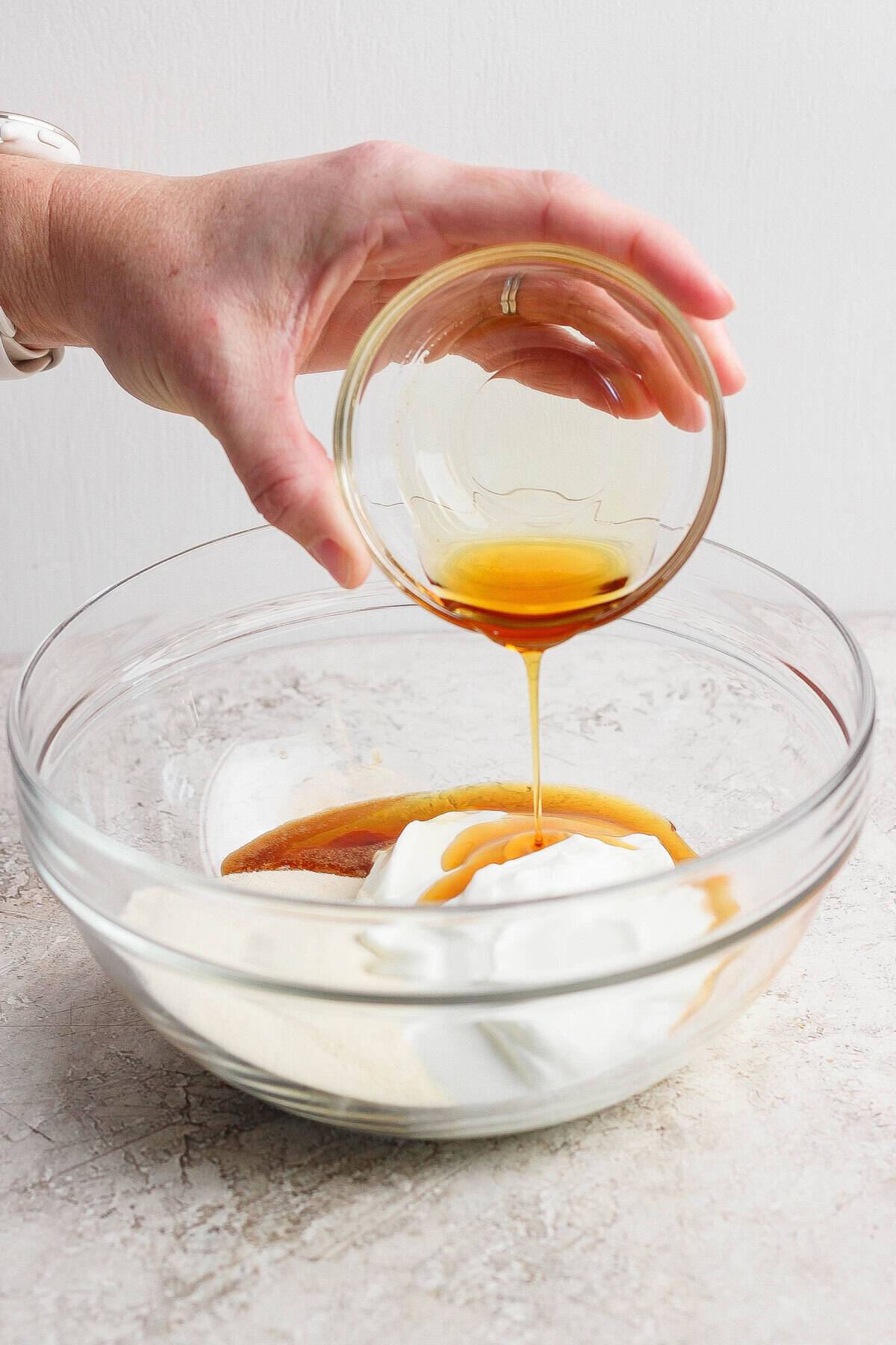 A hand pours a small glass bowl of honey into a larger bowl containing yogurt and other baking ingredients.
