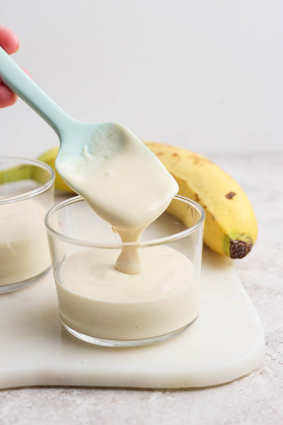 A blue spatula is lifting creamy banana pudding from a glass cup. A ripe banana and another cup of pudding are in the background on a white surface.