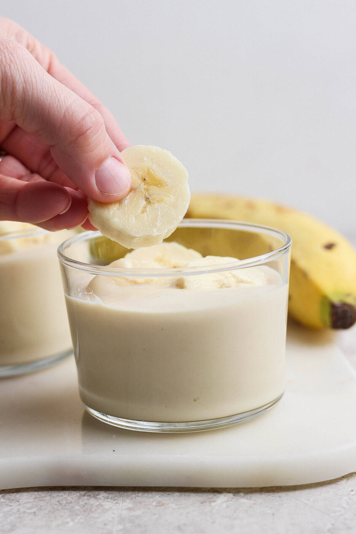 A hand placing a banana slice on top of a glass filled with banana pudding, with a whole banana in the background.
