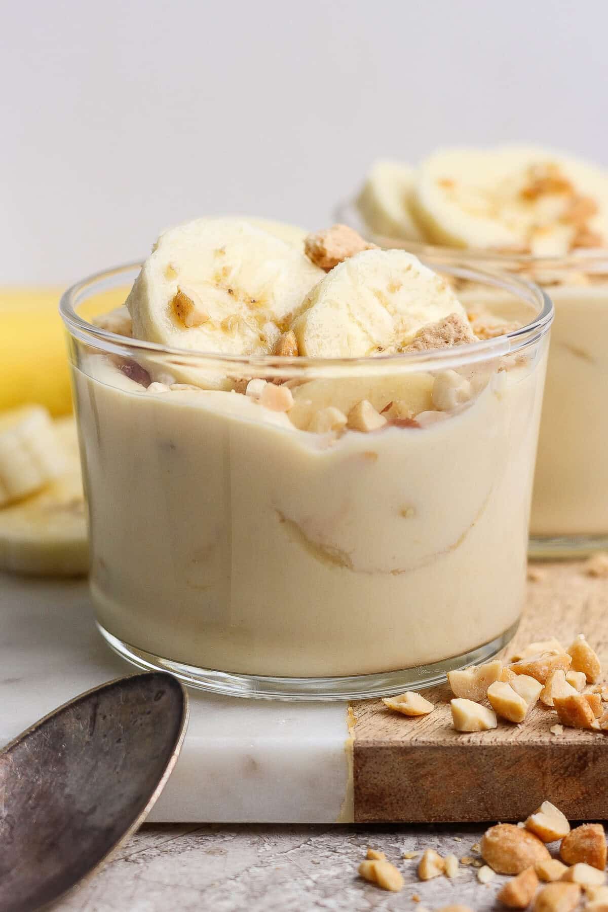 A glass cup filled with banana pudding, topped with banana slices and chopped nuts, placed on a marble surface next to a spoon and more chopped nuts.