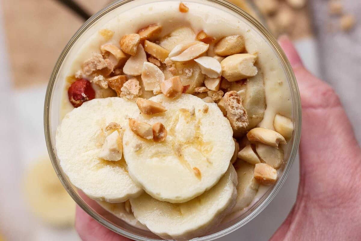 A hand holds a bowl of creamy dessert topped with banana slices and chopped peanuts.