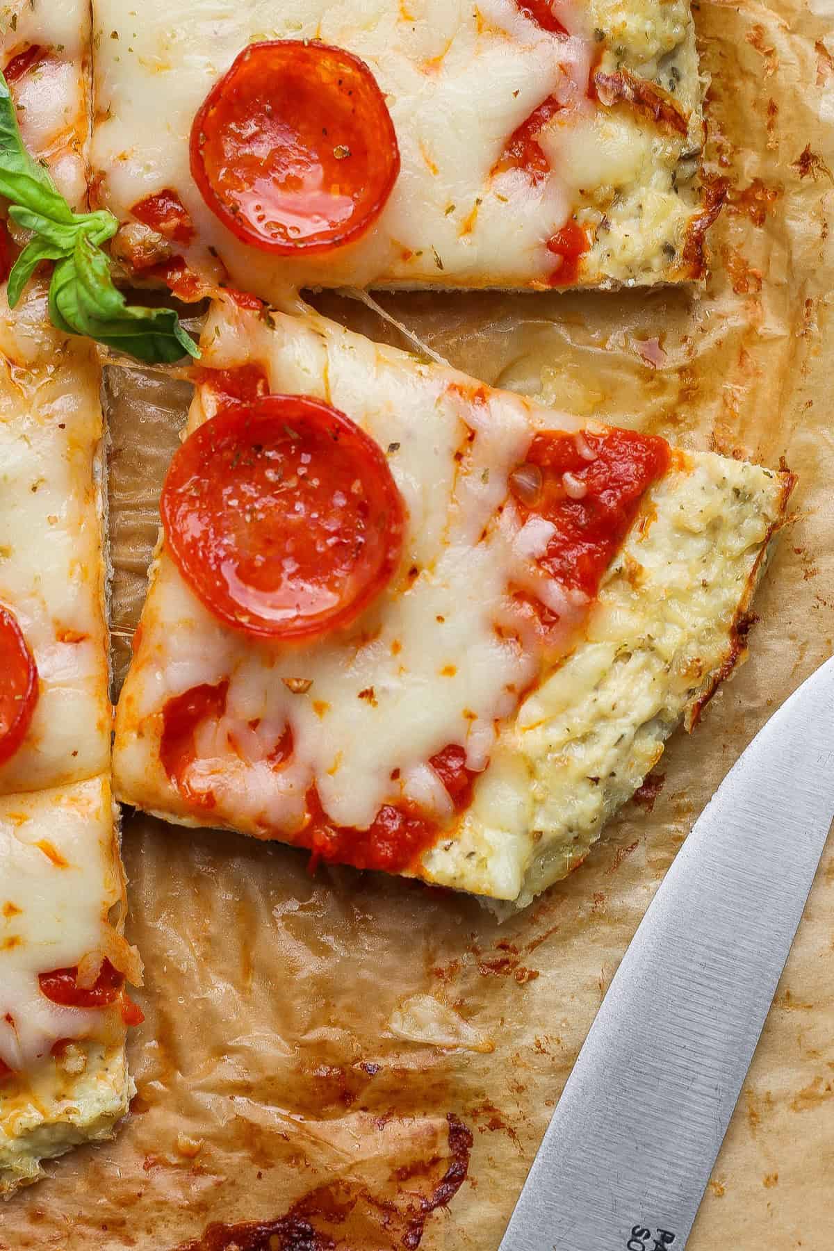 Close-up of a square slice of chicken crust pizza topped with pepperoni and melted cheese on parchment paper, next to a knife.