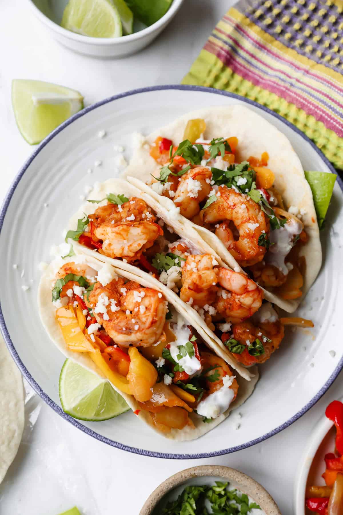 Three shrimp tacos with diced vegetables, cilantro, crumbled cheese, and sauce on flour tortillas, served on a plate with lime wedges.