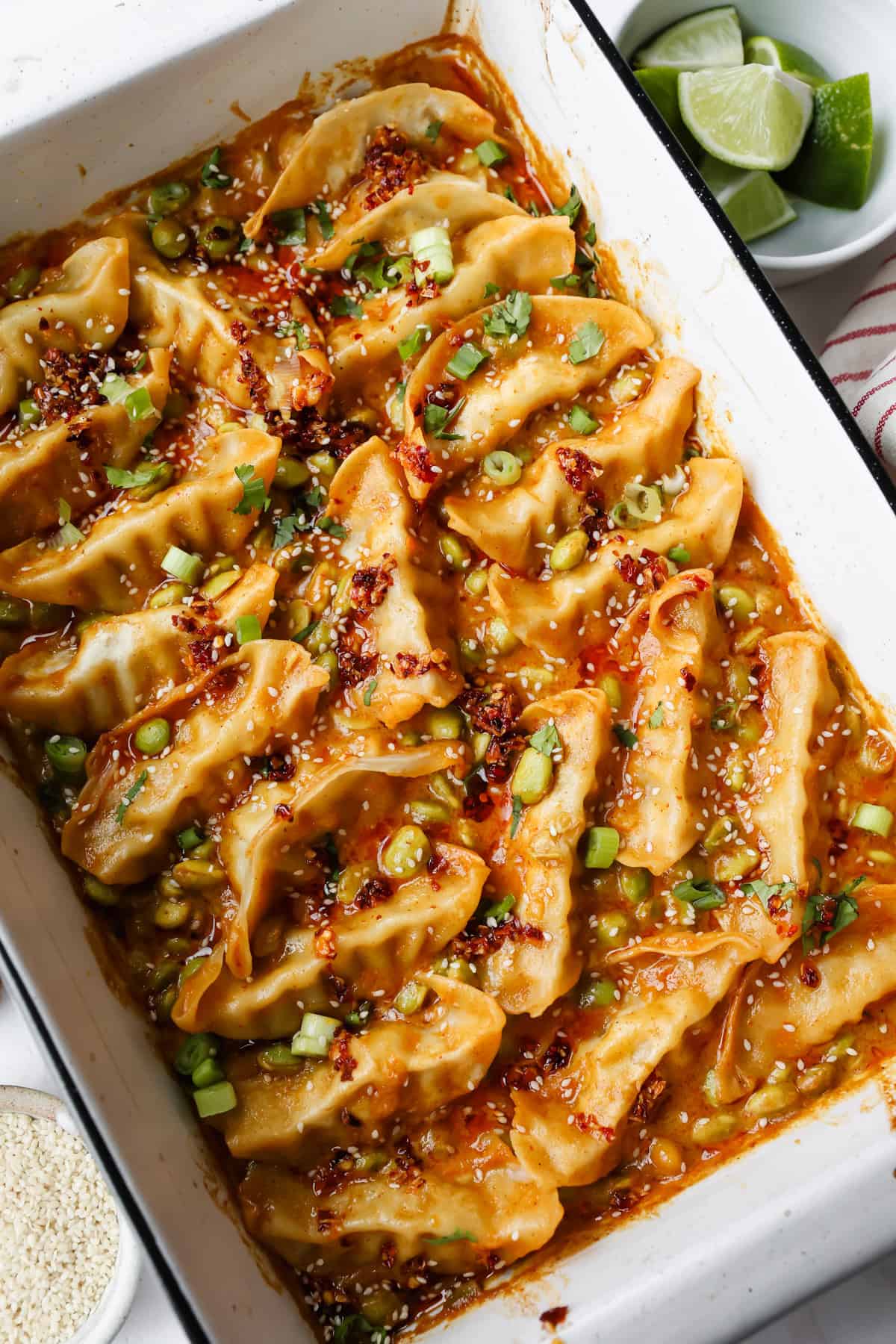 A white baking dish filled with cooked dumplings in a spicy sauce, garnished with chopped green onions, cilantro, and sesame seeds. A plate of lime wedges is nearby.