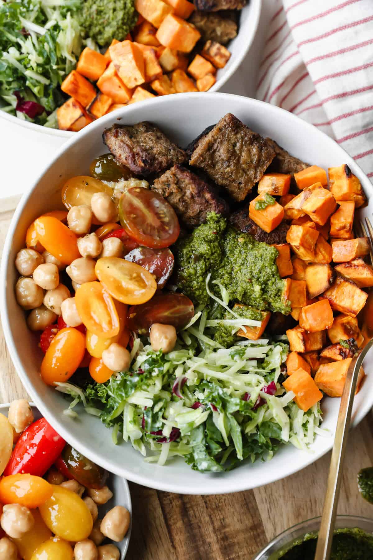 A bowl filled with grilled beef strips, roasted sweet potatoes, cherry tomatoes, chickpeas, shredded greens, and green sauce, with a fork and another bowl partially visible.