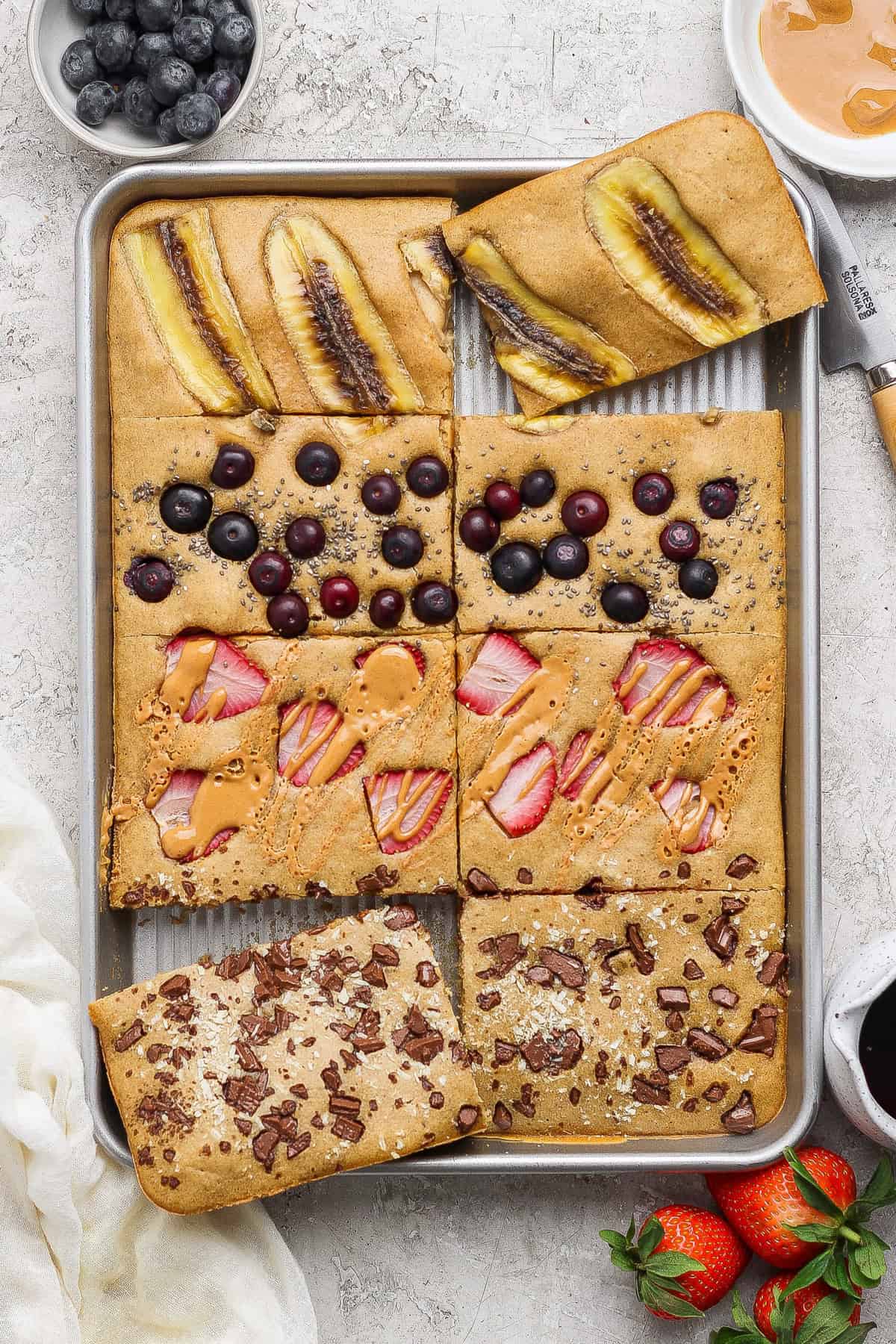 A baking tray with six rectangular pieces of sheet pancakes, each topped with different ingredients: banana, blueberries, strawberries with peanut butter, and chocolate chunks.