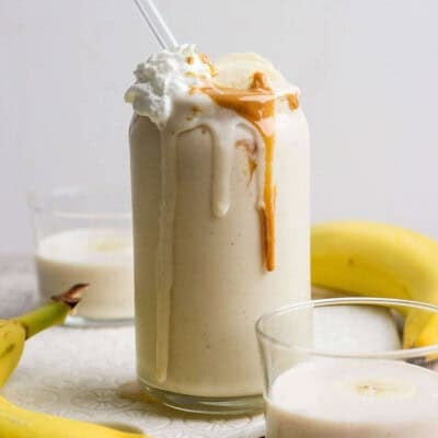 A glass of peanut butter banana protein shake topped with whipped cream, banana slices, and peanut butter, with a straw, surrounded by bananas and two smaller glasses of smoothie.