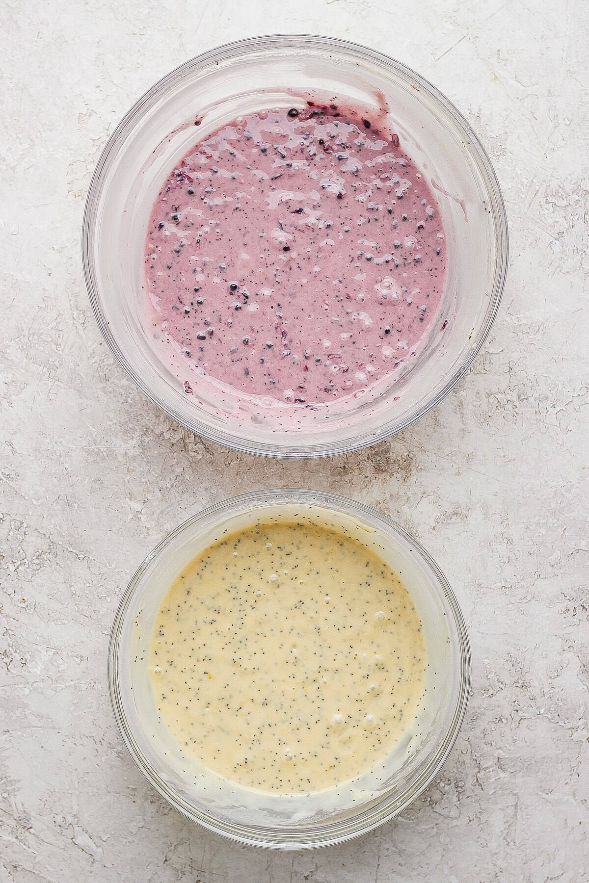 Two glass bowls on a light surface, one with purple berry-speckled batter and one with yellow batter containing small black seeds.