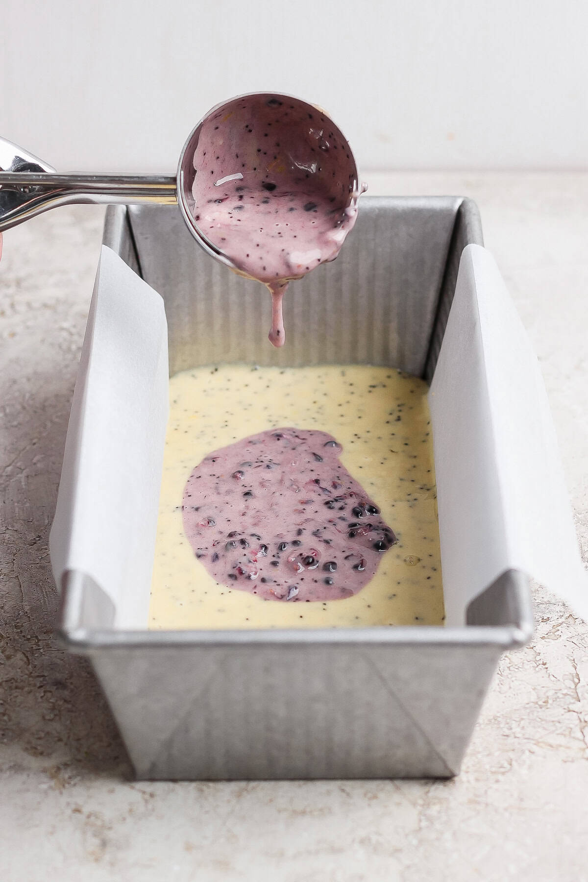 A metal scoop pours purple berry batter over yellow batter in a parchment-lined loaf pan on a light surface.