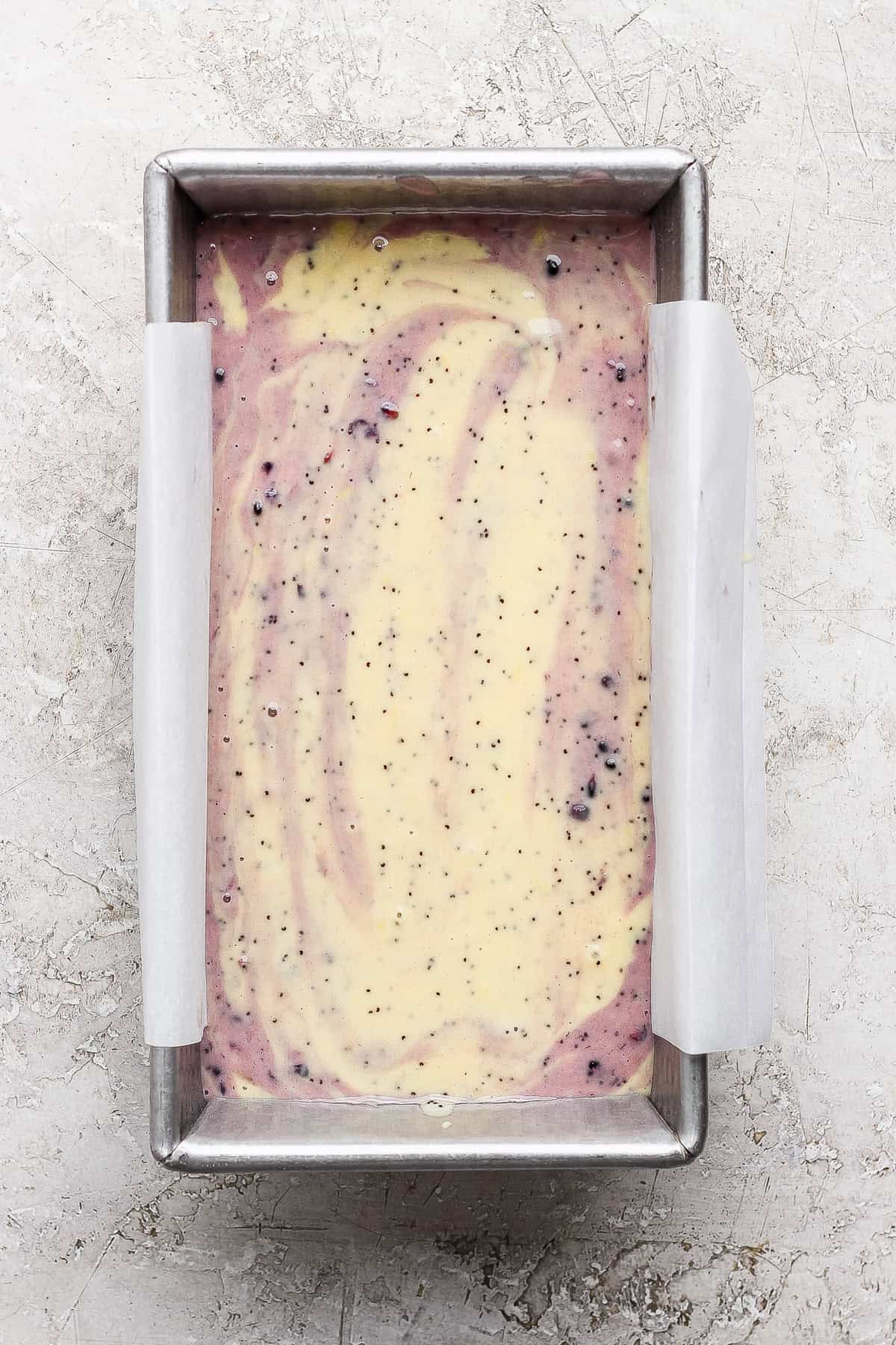 Rectangular loaf pan lined with parchment paper, filled with swirled yellow and purple cake batter containing small dark specks, on a light textured surface.