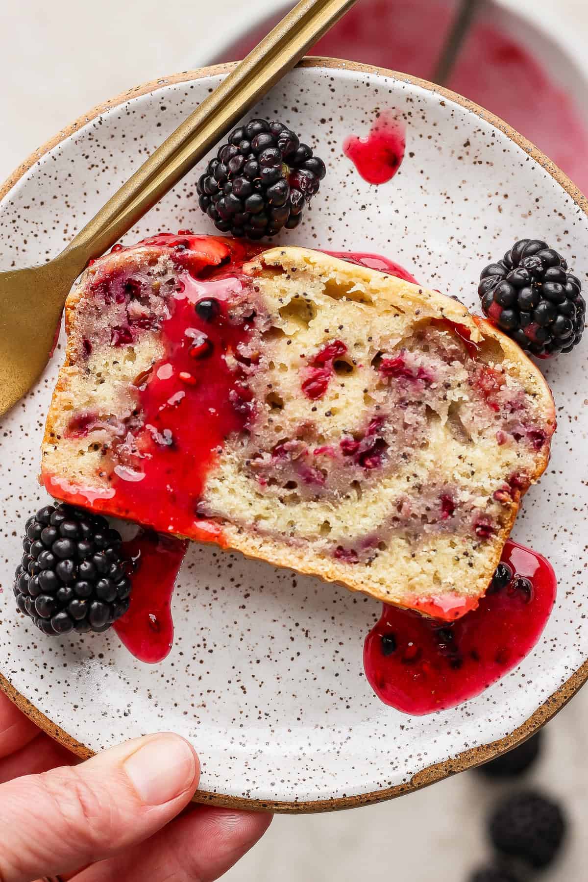 A slice of berry loaf cake with berry sauce and fresh blackberries on a speckled plate, with a gold fork beside it.