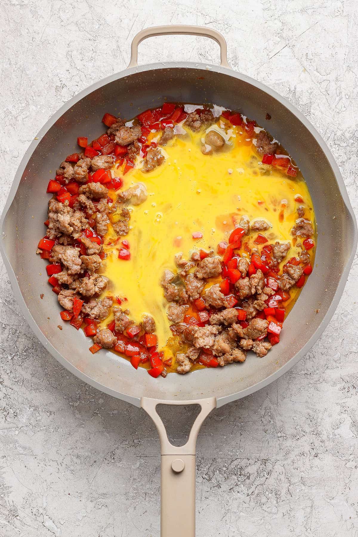 A skillet containing cooked sausage, diced red bell peppers, and beaten eggs beginning to cook.
