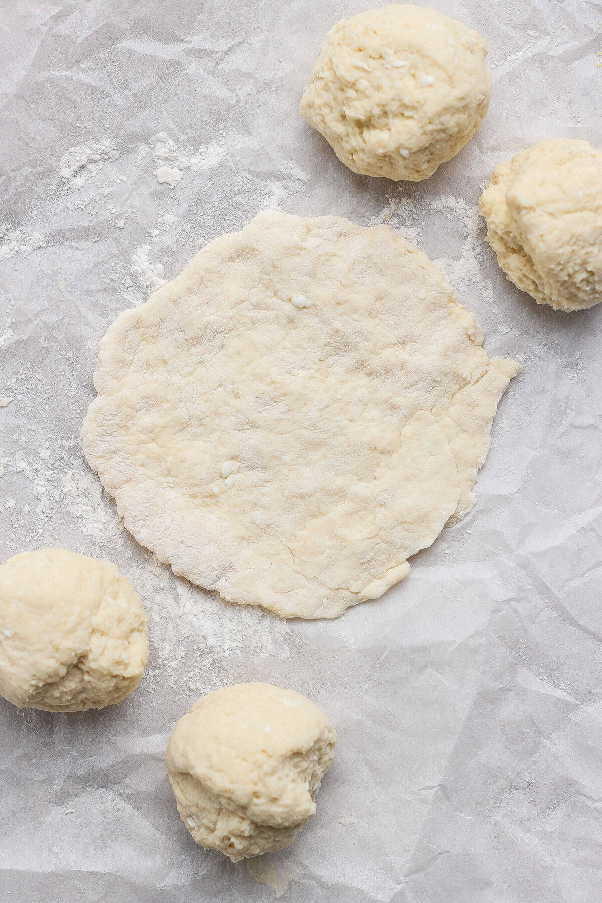 Four dough balls and one flattened round of dough sit on a sheet of parchment paper dusted with flour.