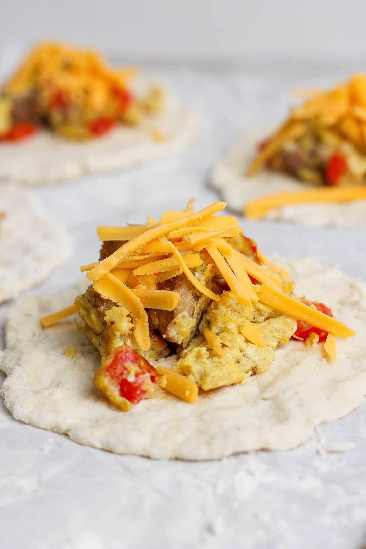 Uncooked dough topped with scrambled eggs, red bell peppers, and shredded cheddar cheese, ready to be assembled.