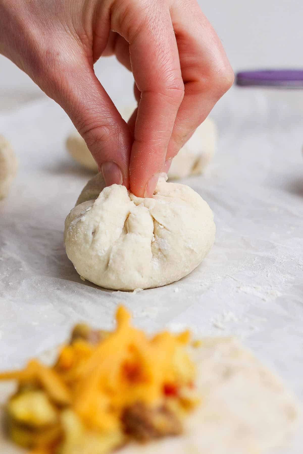 A hand pinches the top of a ball of dough to seal it closed; fillings and shredded cheese are visible on a piece of dough in the foreground.