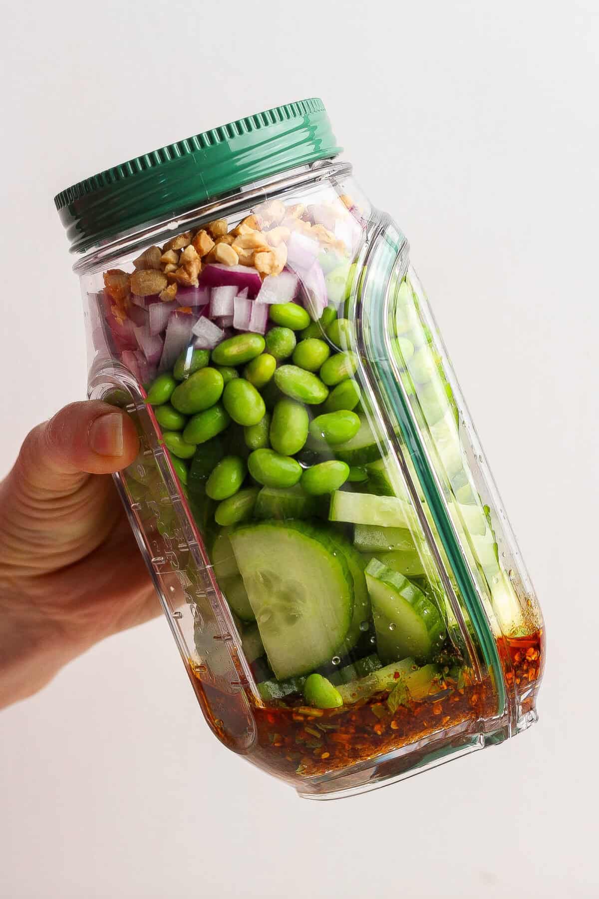 A hand holds a tilted plastic jar filled with layered ingredients including cucumber, edamame, red onion, walnuts, and dressing at the bottom.