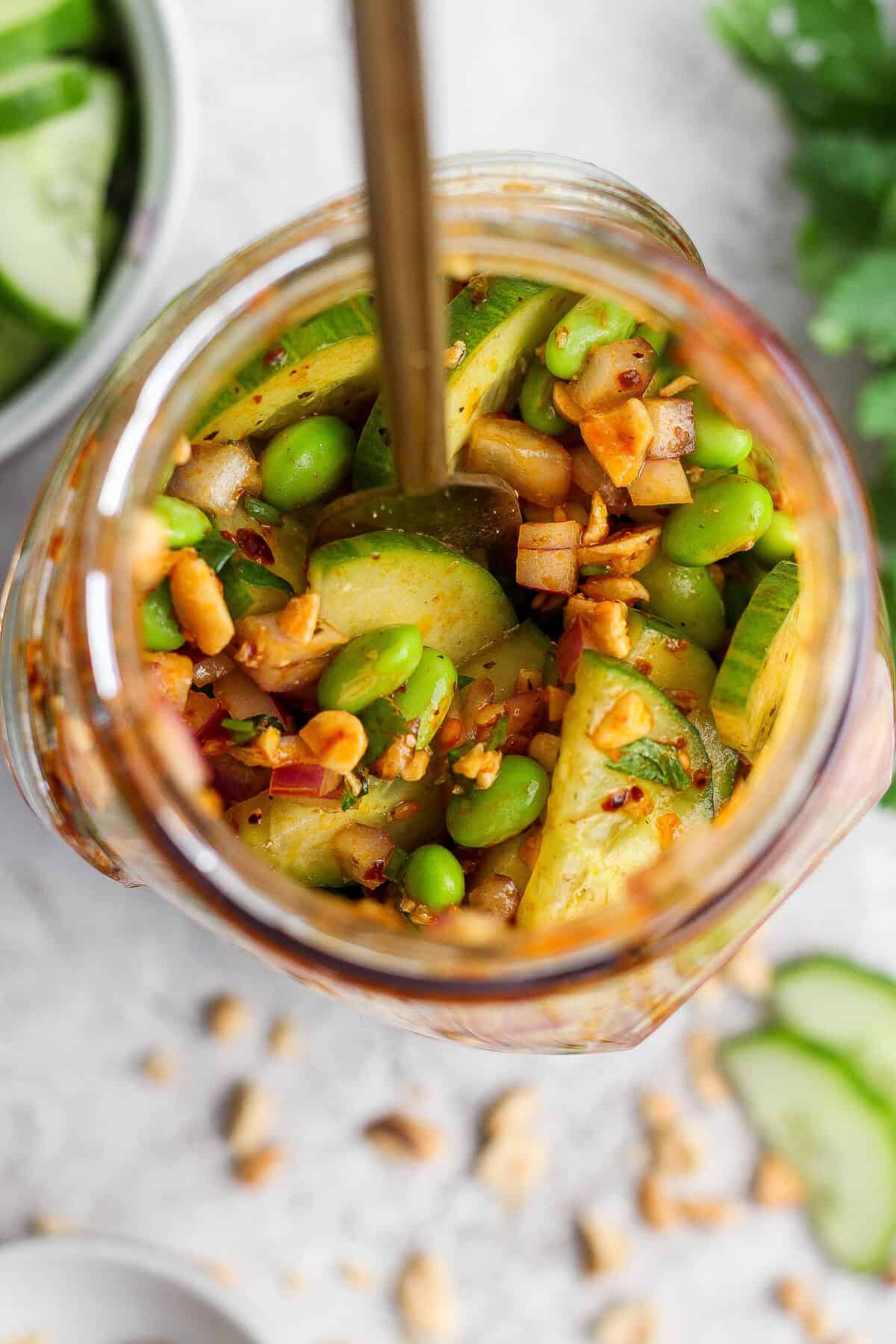Jar filled with a mix of sliced cucumbers, edamame, chopped red onions, peanuts, and spices, with a spoon inside and some ingredients scattered around.