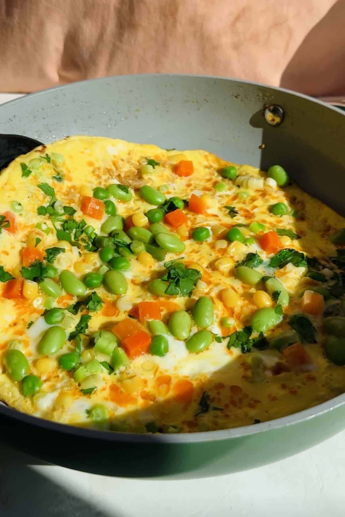 Omelette in a pan topped with chopped carrots, edamame, corn, and fresh herbs, shown in natural sunlight.