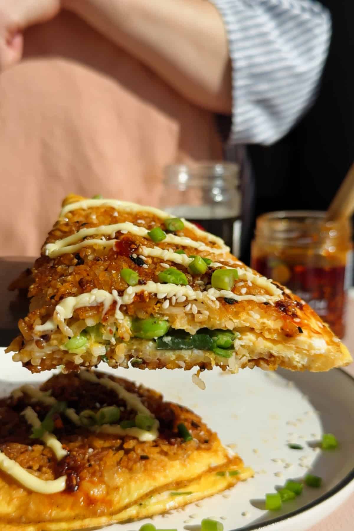 A close-up of a savory slice of food topped with chopped green onions and a drizzle of mayonnaise, being held above a plate with more slices.