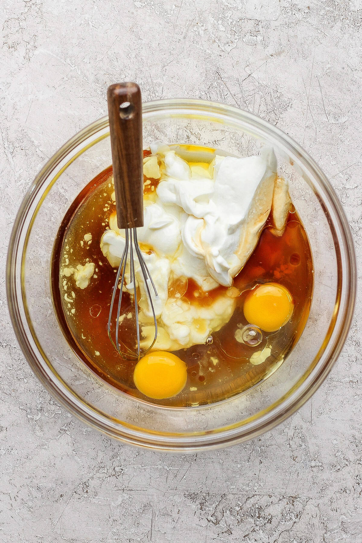 A glass bowl containing two raw eggs, yogurt, and a brown liquid with a metal whisk resting on the side, placed on a textured countertop.
