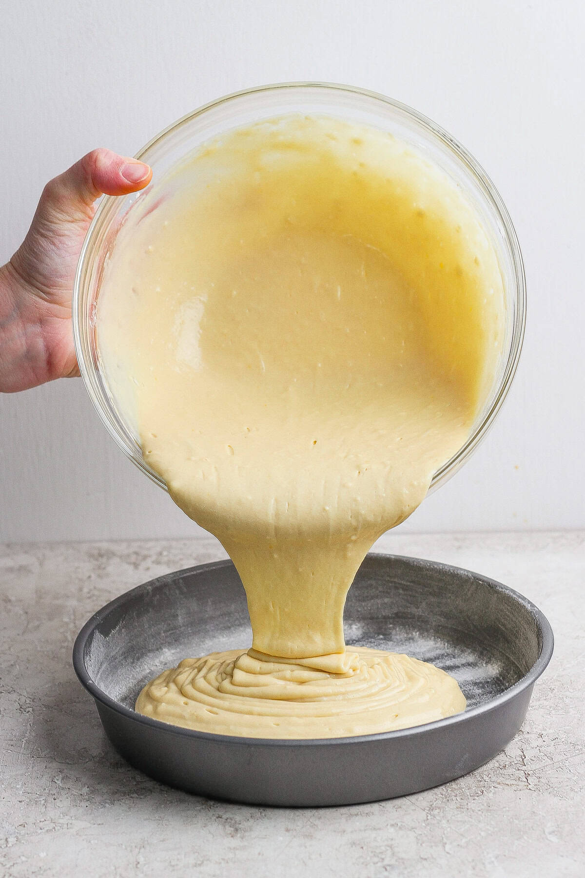 A hand pours cake batter from a glass bowl into a round, floured metal baking pan on a light countertop.