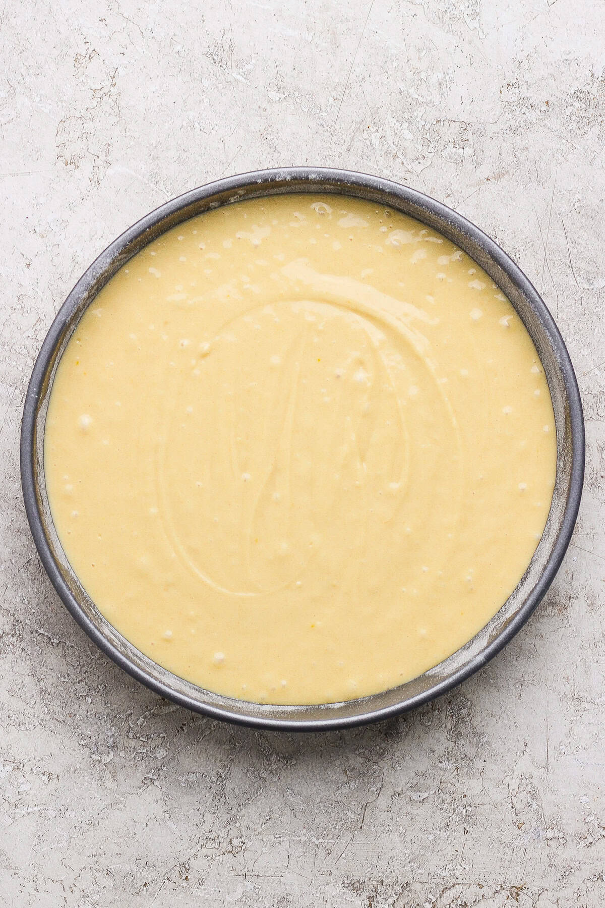 A round cake pan filled with smooth, pale yellow cake batter sits on a light textured surface.