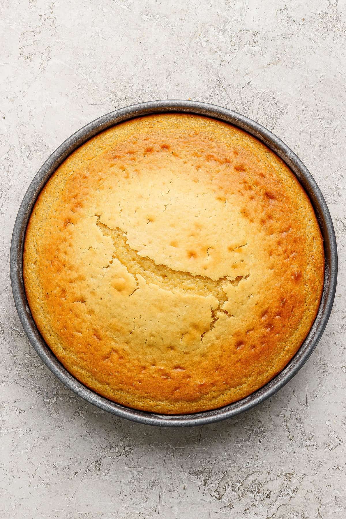 A round, golden-brown cake with a crack across the center sits in a metal baking pan on a light textured surface.
