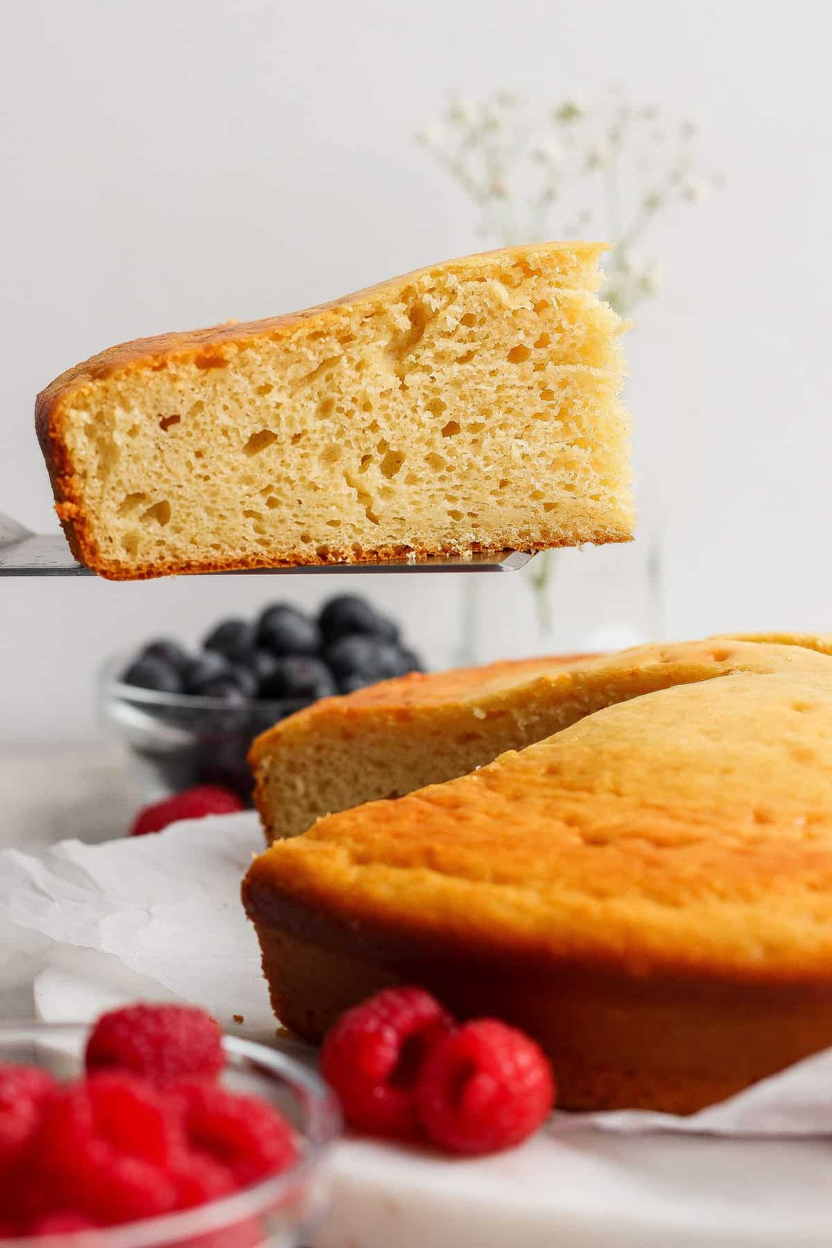 A slice of plain yellow cake being lifted from a round cake, with fresh raspberries and blueberries in bowls nearby.