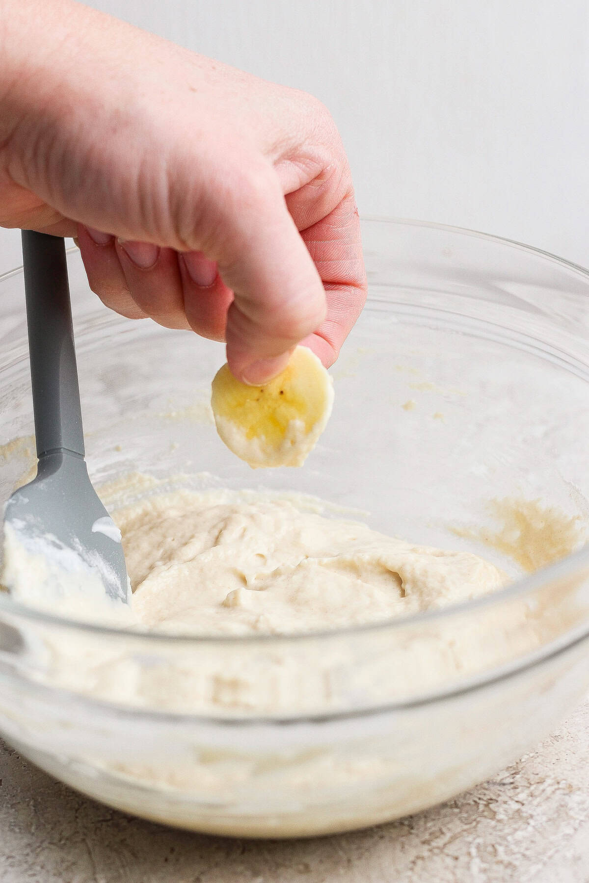 A hand holds a banana slice above a bowl of batter with a spatula resting inside the bowl.