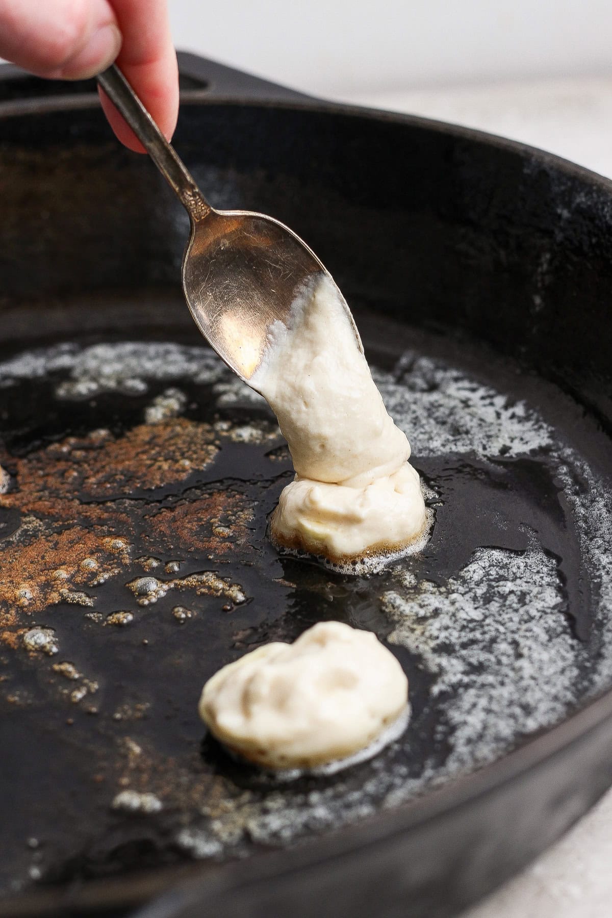 A hand uses a spoon to drop batter into a black skillet with melted butter.