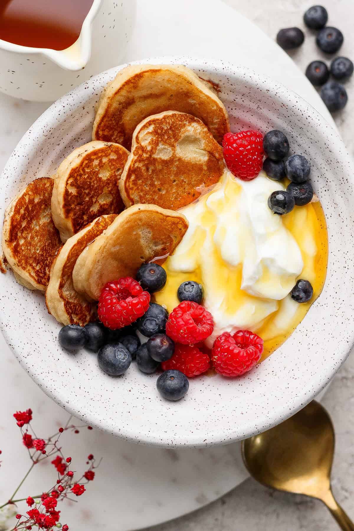 A bowl with small pancakes, yogurt, raspberries, blueberries, and honey, placed on a white plate with extra berries, a gold spoon, and a small pitcher of syrup nearby.