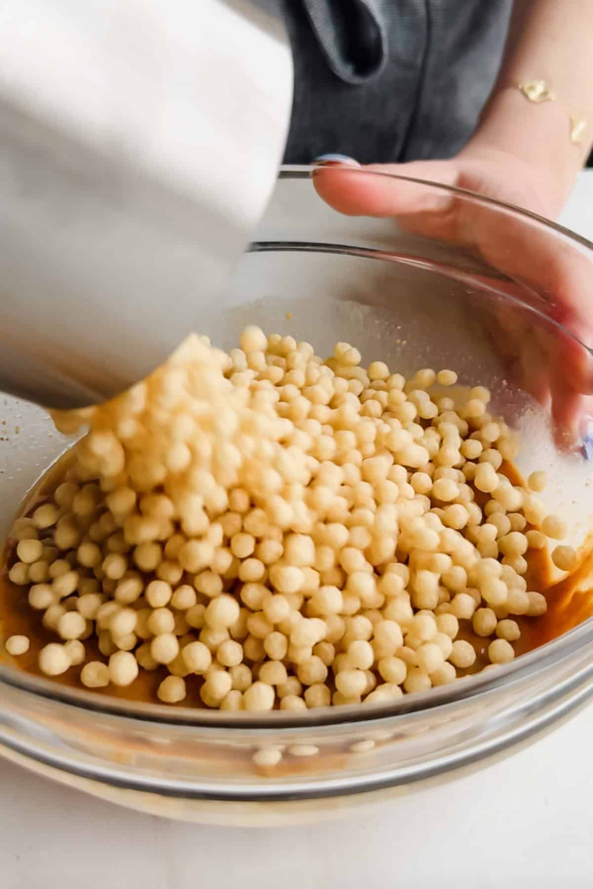 A person pours small round cereal pieces from a metal container into a glass bowl containing a brown liquid mixture.