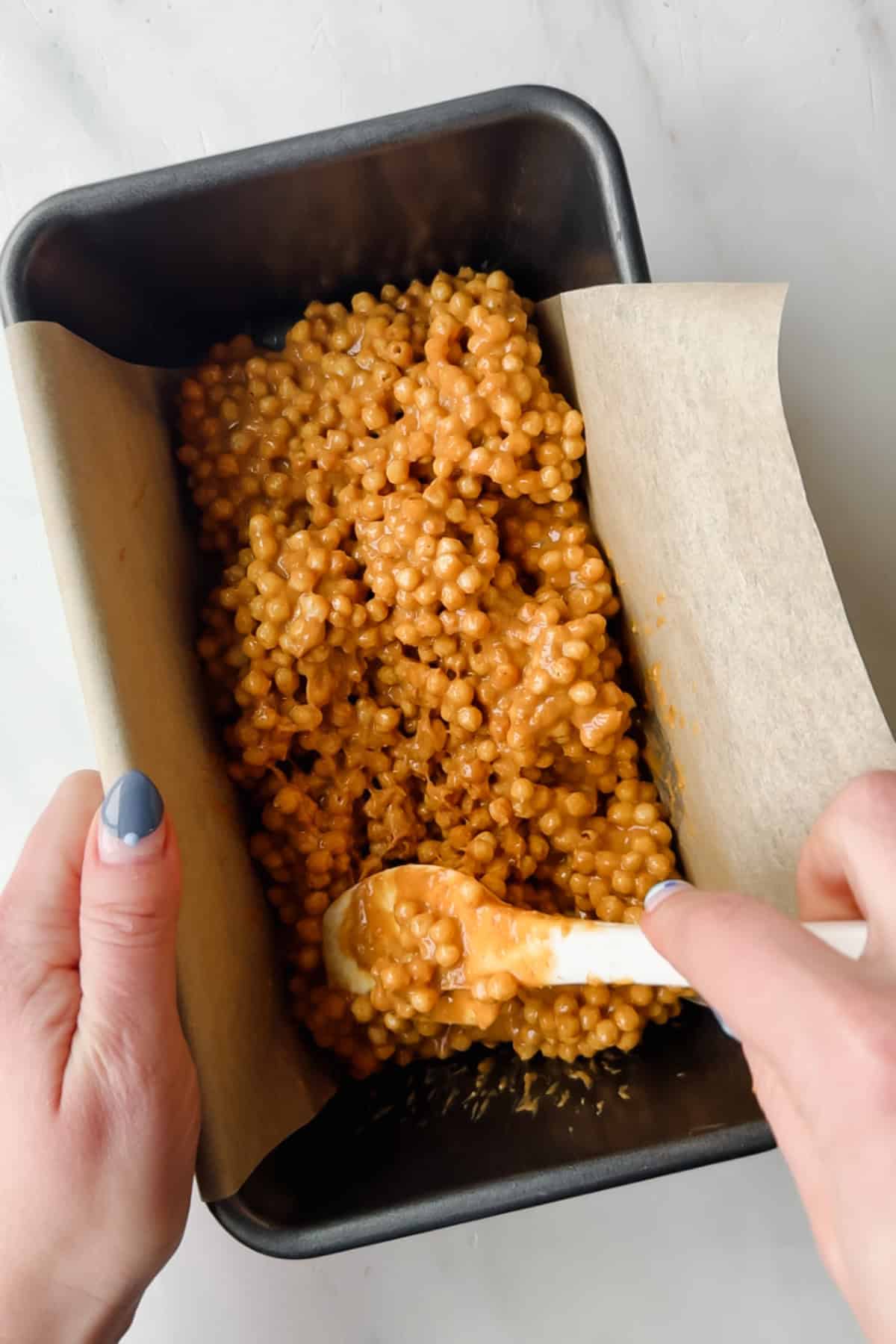 Hands spreading a mixture of coated puffed cereal into a parchment-lined loaf pan with a white spatula.