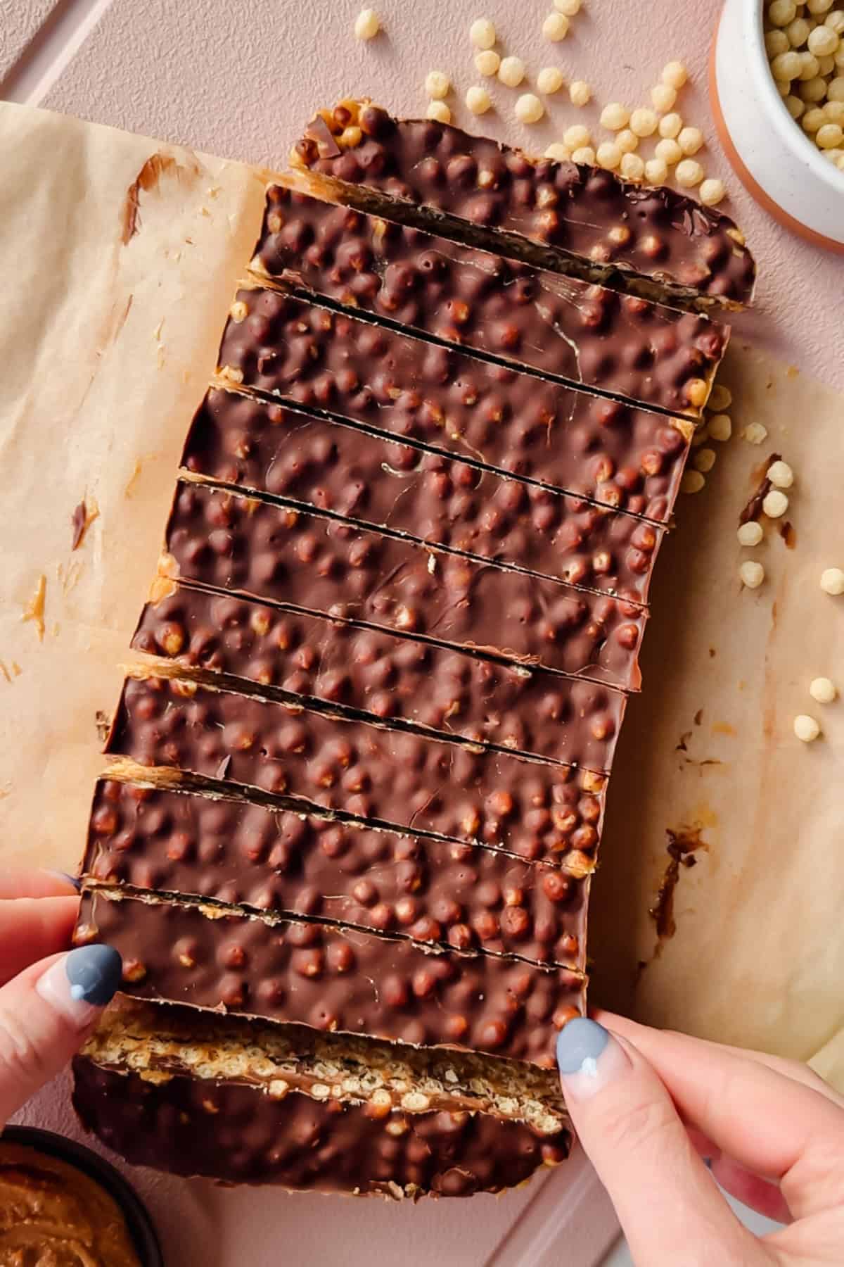 Rectangular chocolate-covered crispy bars with a textured surface, sliced into strips on parchment paper. A person holds one end; puffed grains and a bowl are nearby.