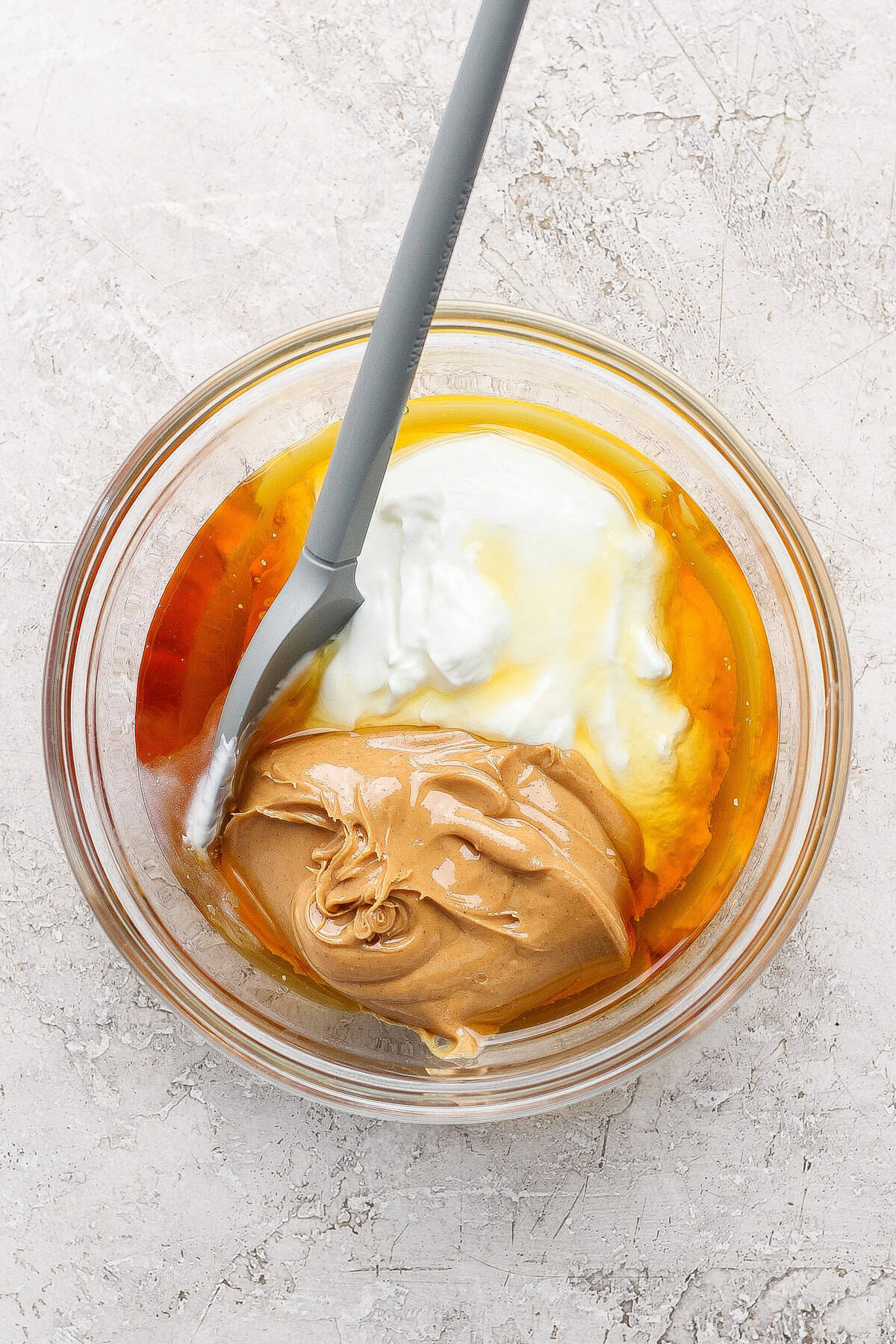 A glass bowl containing peanut butter, yogurt, honey, and an egg with a gray spatula resting inside.