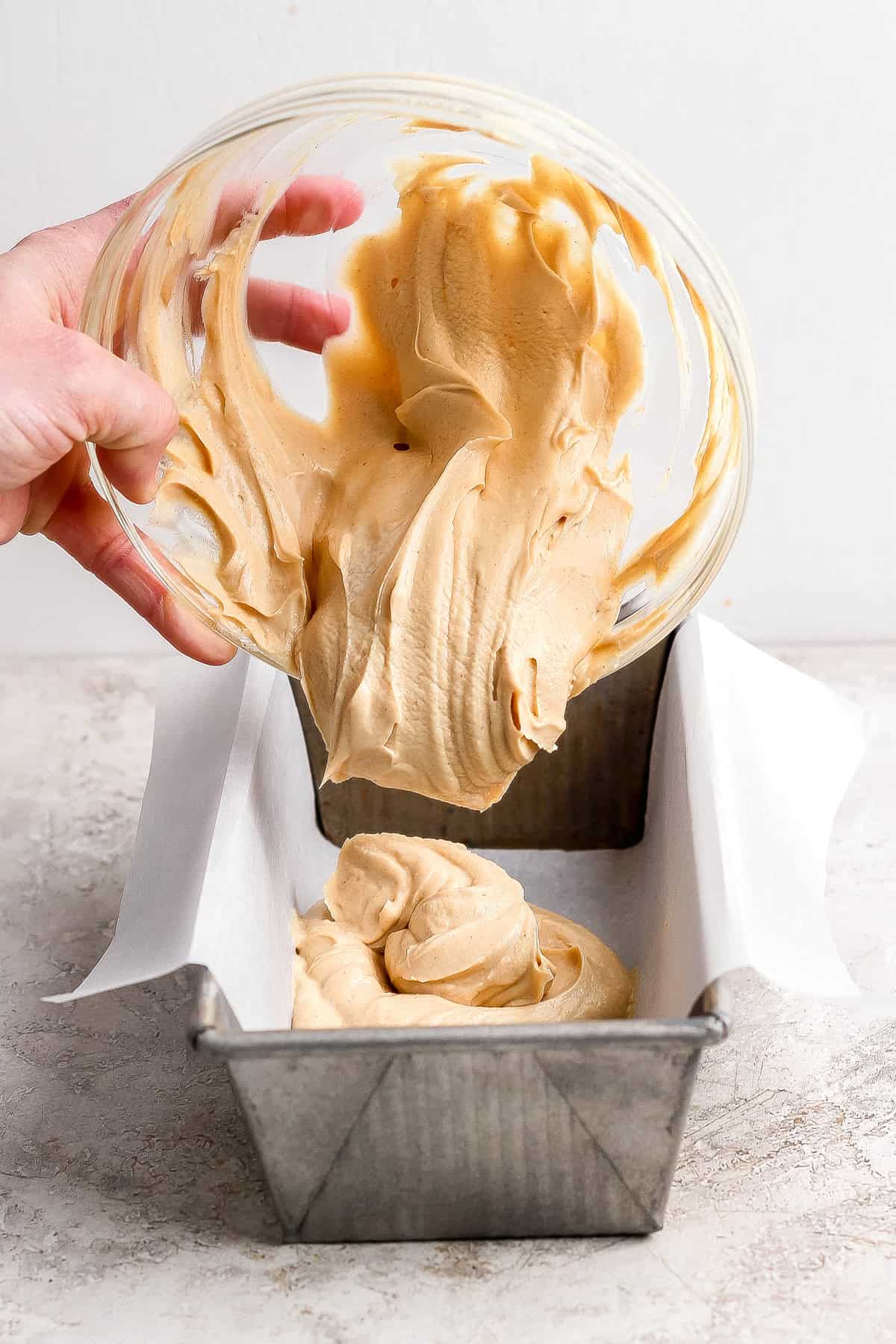 A person pours thick, light brown batter from a glass bowl into a parchment-lined metal loaf pan on a light-colored surface.