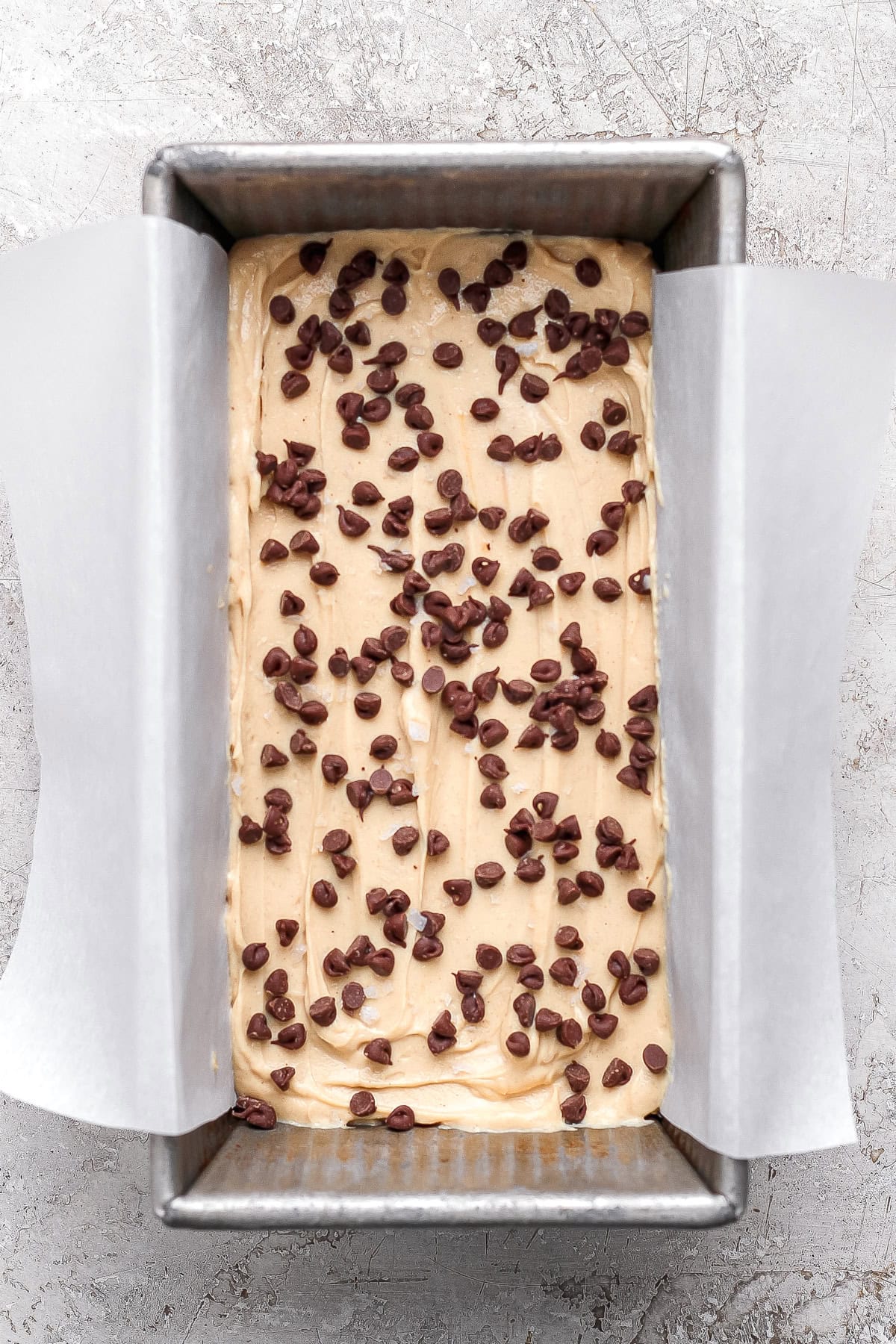 A rectangular metal loaf pan lined with parchment paper, filled with batter and topped with scattered chocolate chips, ready to be baked.