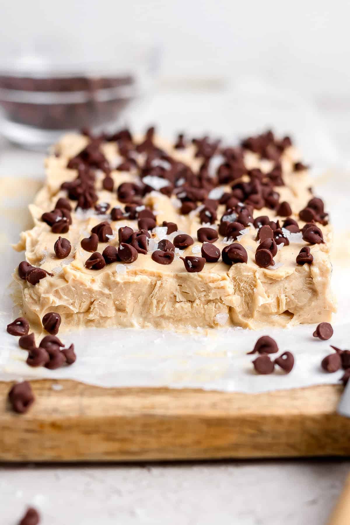 A slab of peanut butter fudge topped with chocolate chips sits on parchment paper on a wooden board.