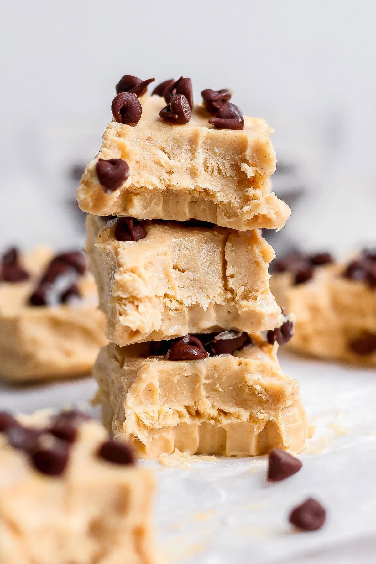 Three pieces of peanut butter fudge, stacked and topped with chocolate chips, with more pieces and chips scattered in the background.