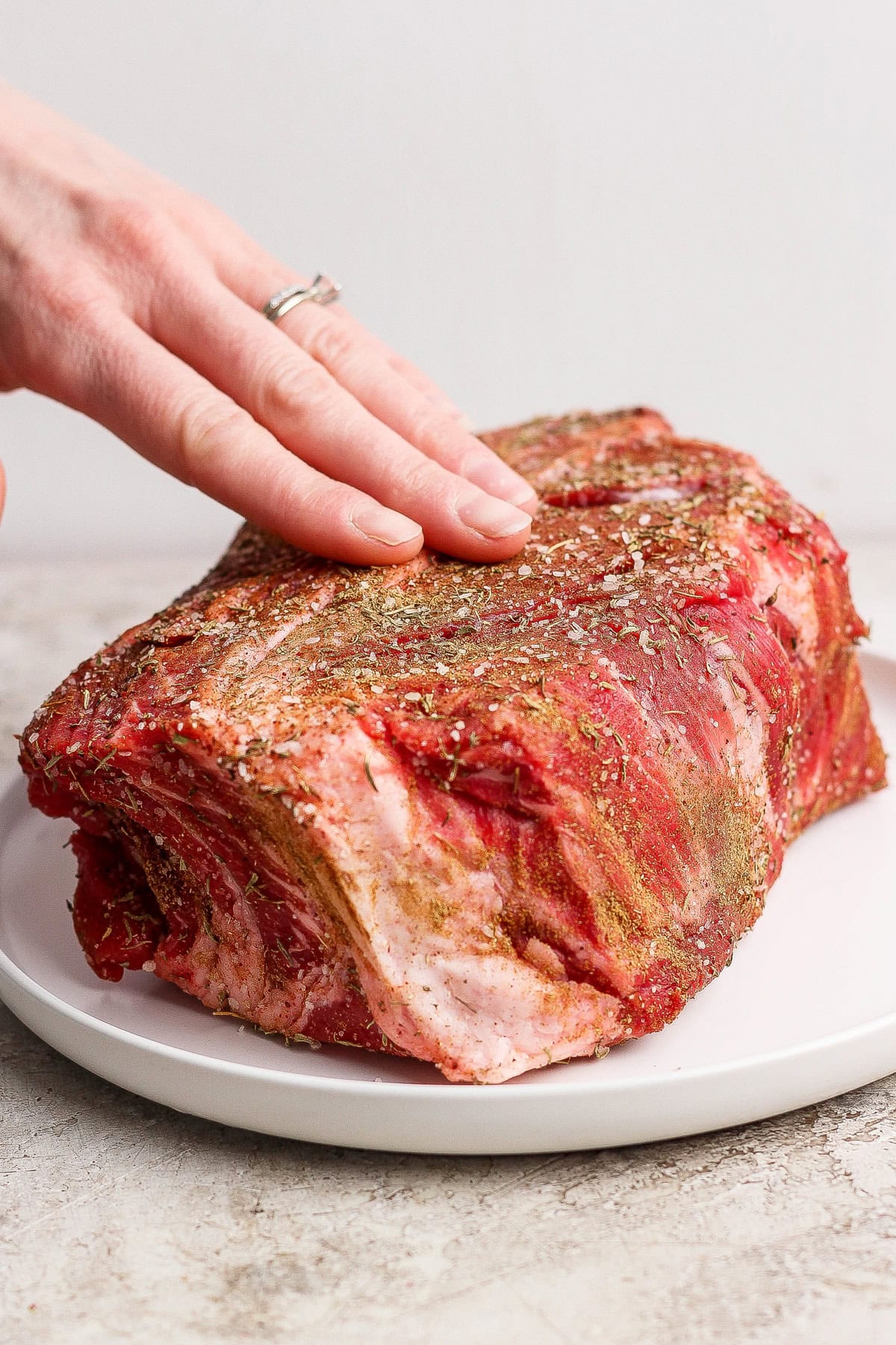 A hand seasoning a raw beef roast with spices and herbs on a white plate.
