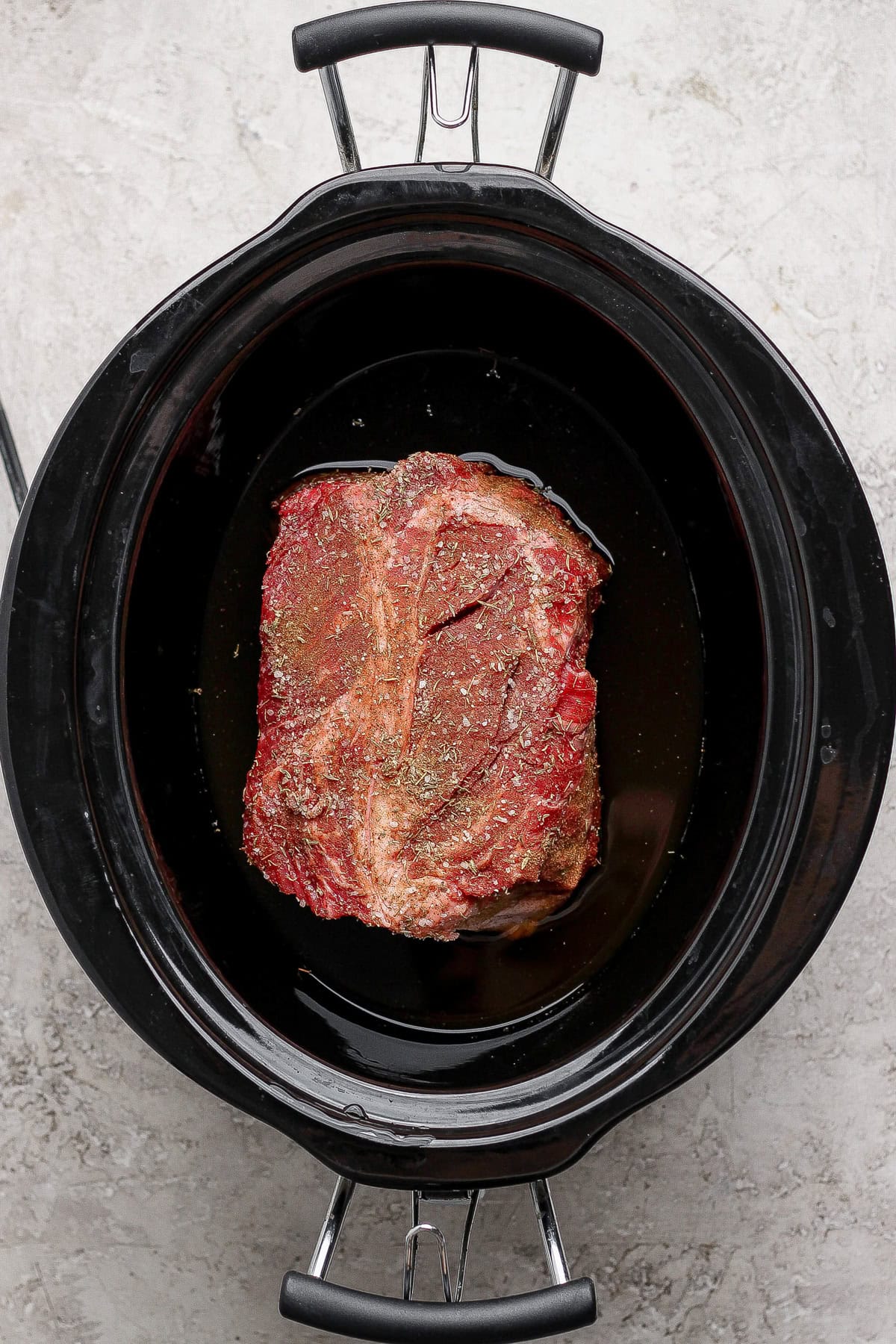 Raw seasoned beef roast sits in a black slow cooker with some liquid at the bottom, viewed from above on a light-colored surface.