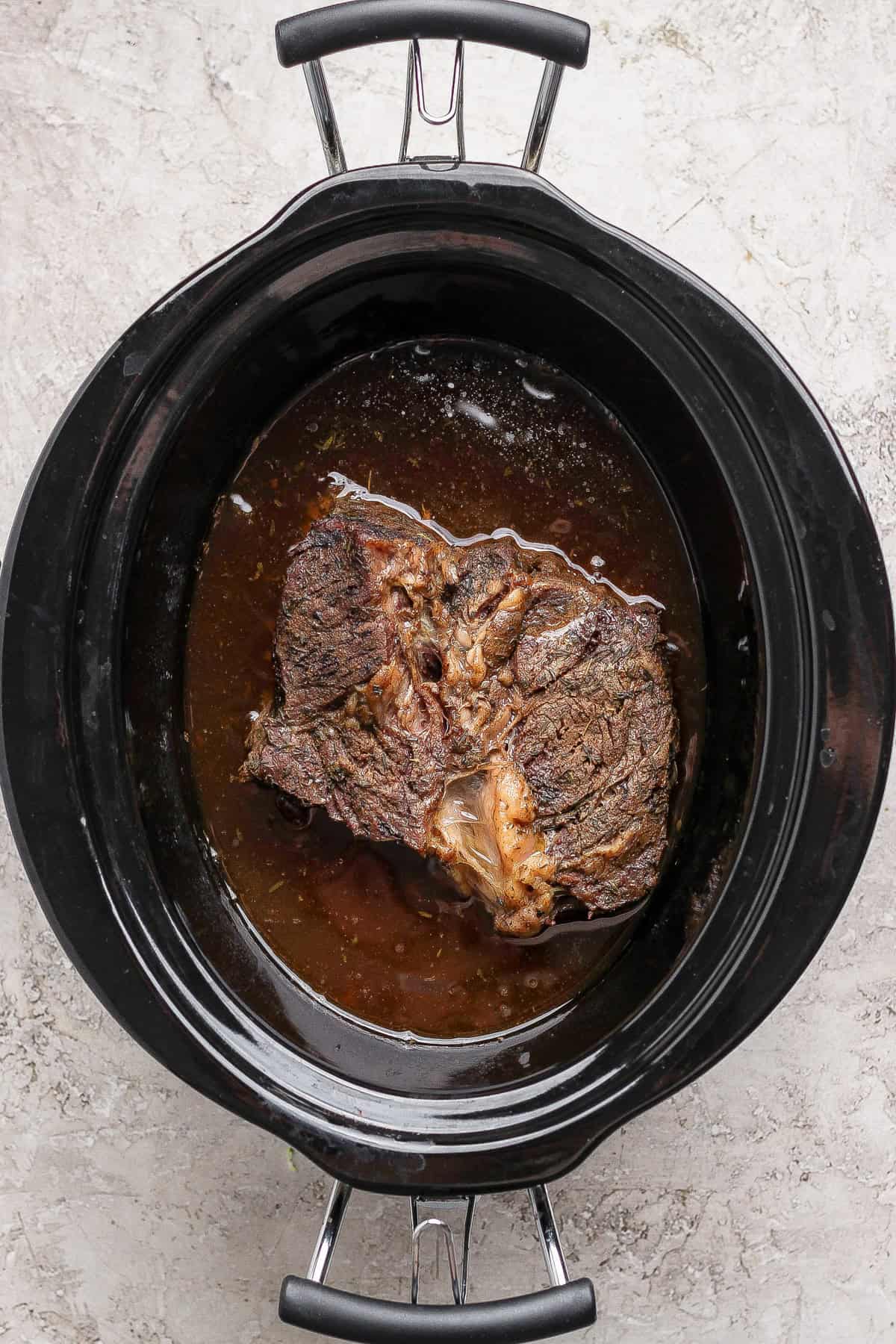 A cooked beef roast sits in broth inside a black slow cooker on a light-colored countertop.