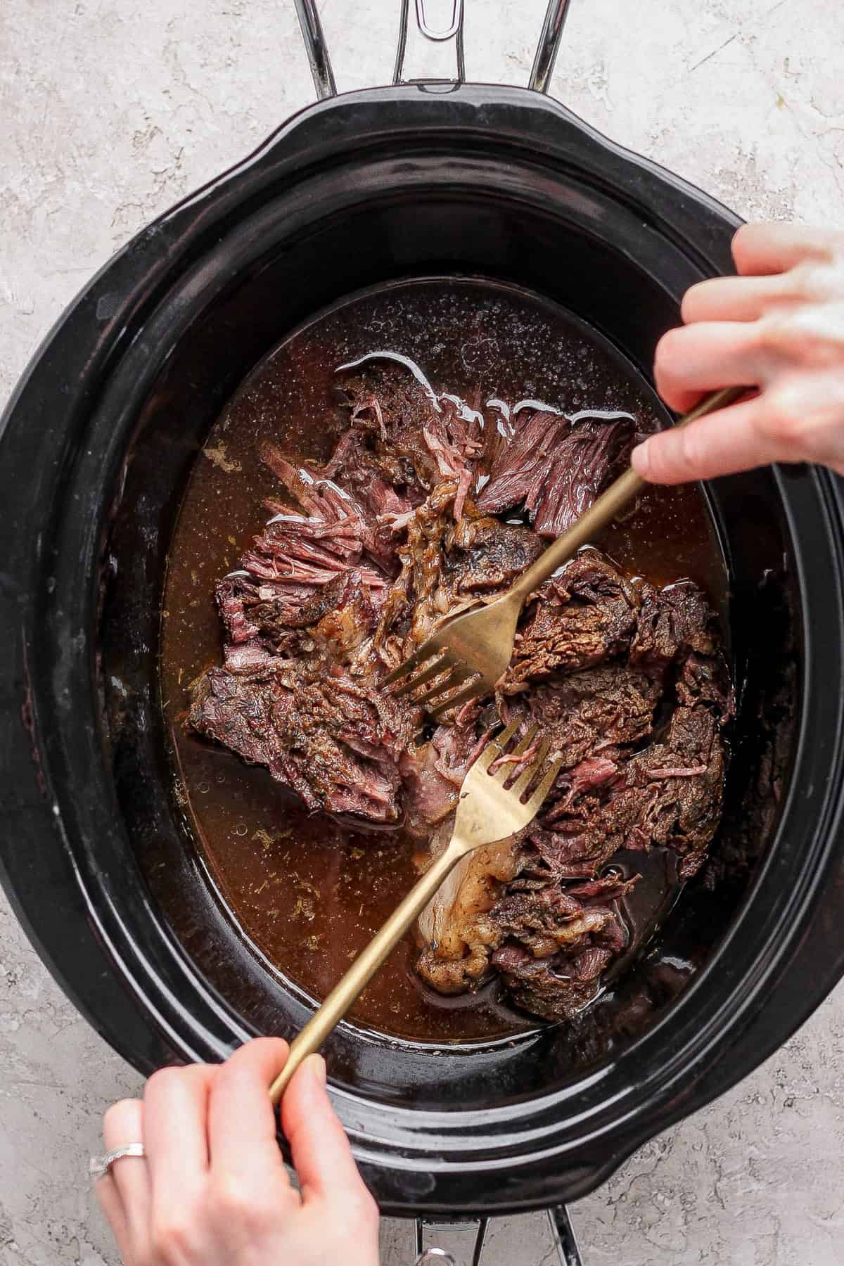 Two hands use forks to shred cooked beef inside a black slow cooker filled with broth on a light countertop.