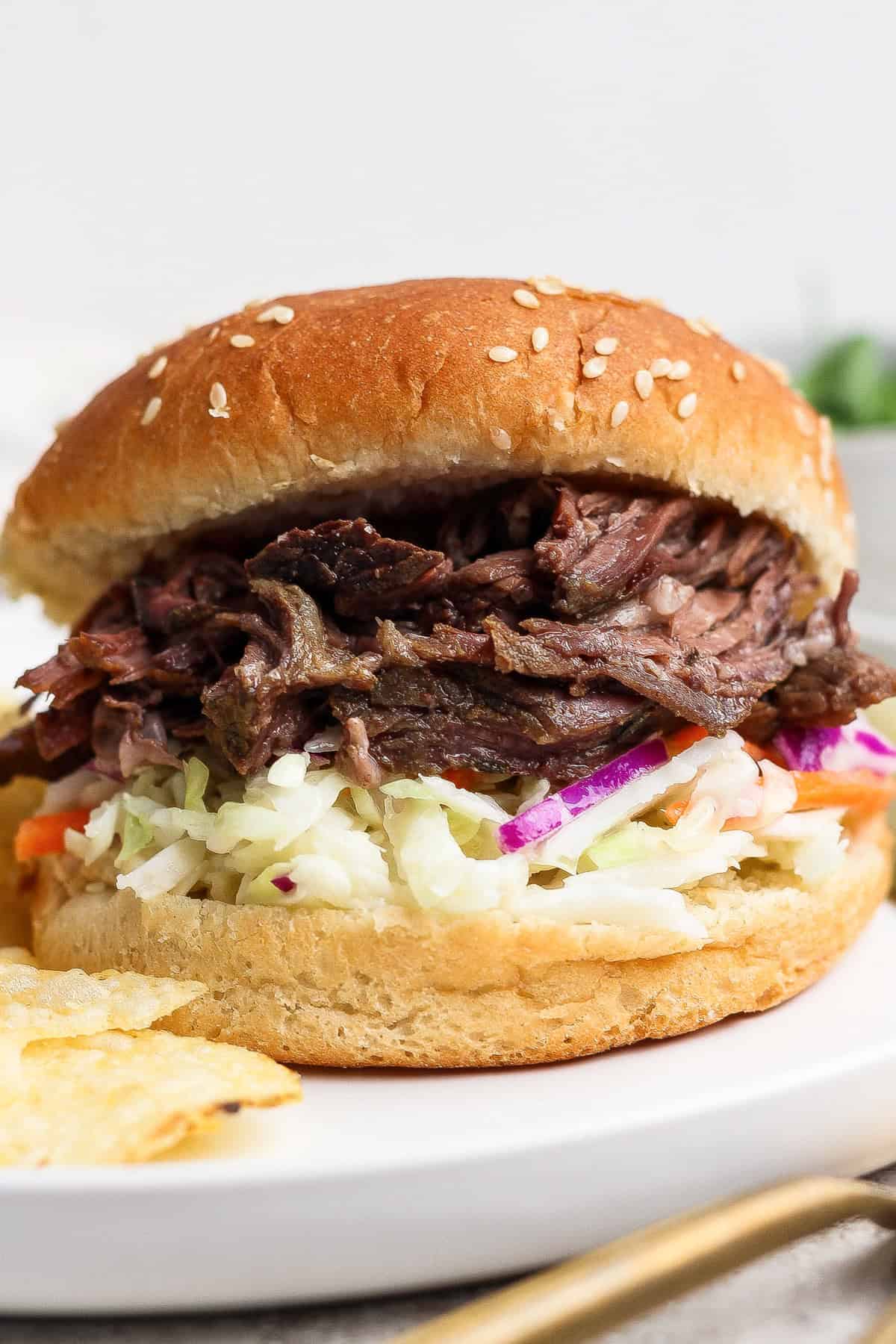 A pulled beef sandwich with coleslaw on a sesame seed bun is served on a white plate with some potato chips.