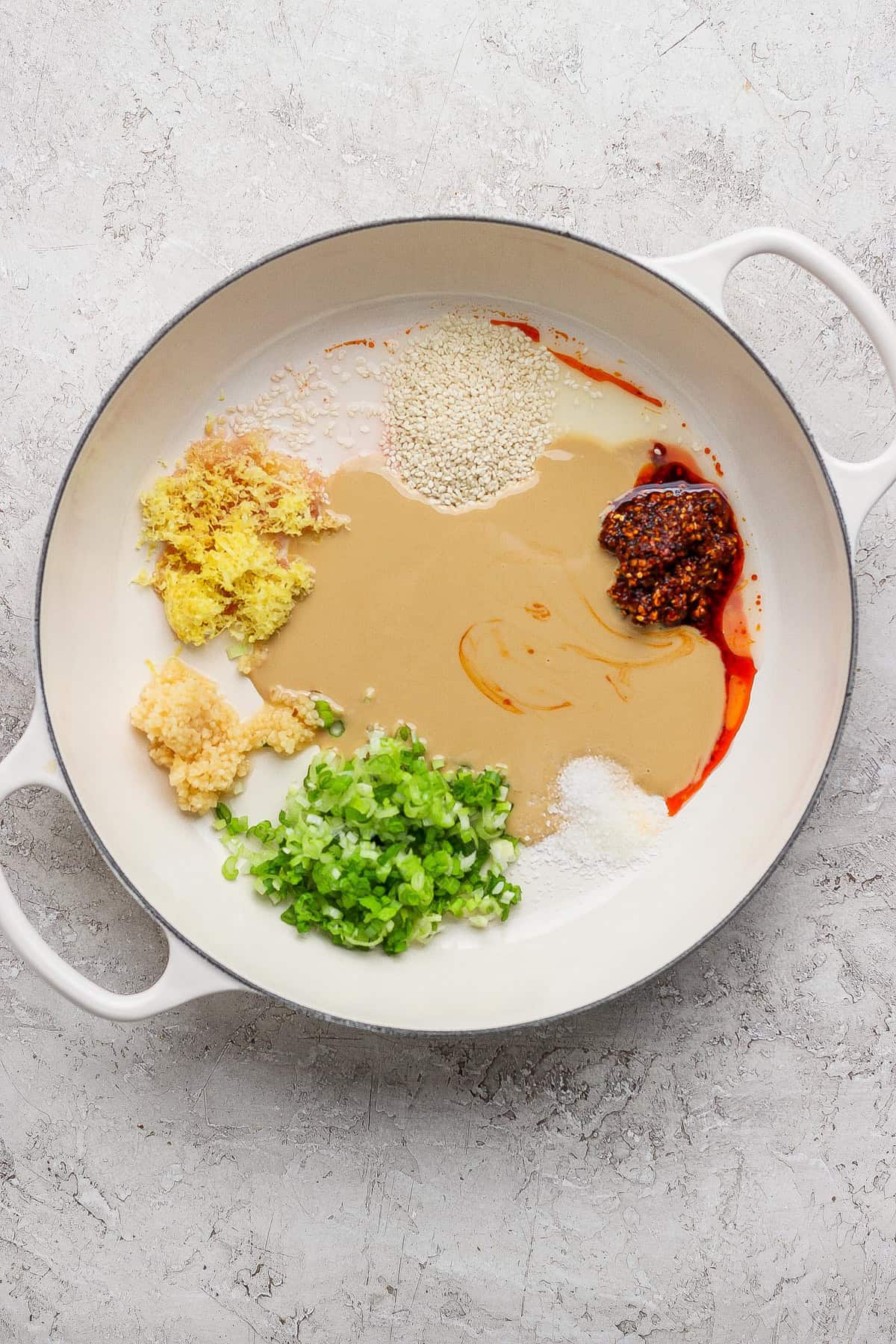 A white pan contains green onions, minced garlic, ginger, sesame seeds, tahini, chili oil, whole grain mustard, and sugar on a light textured surface.