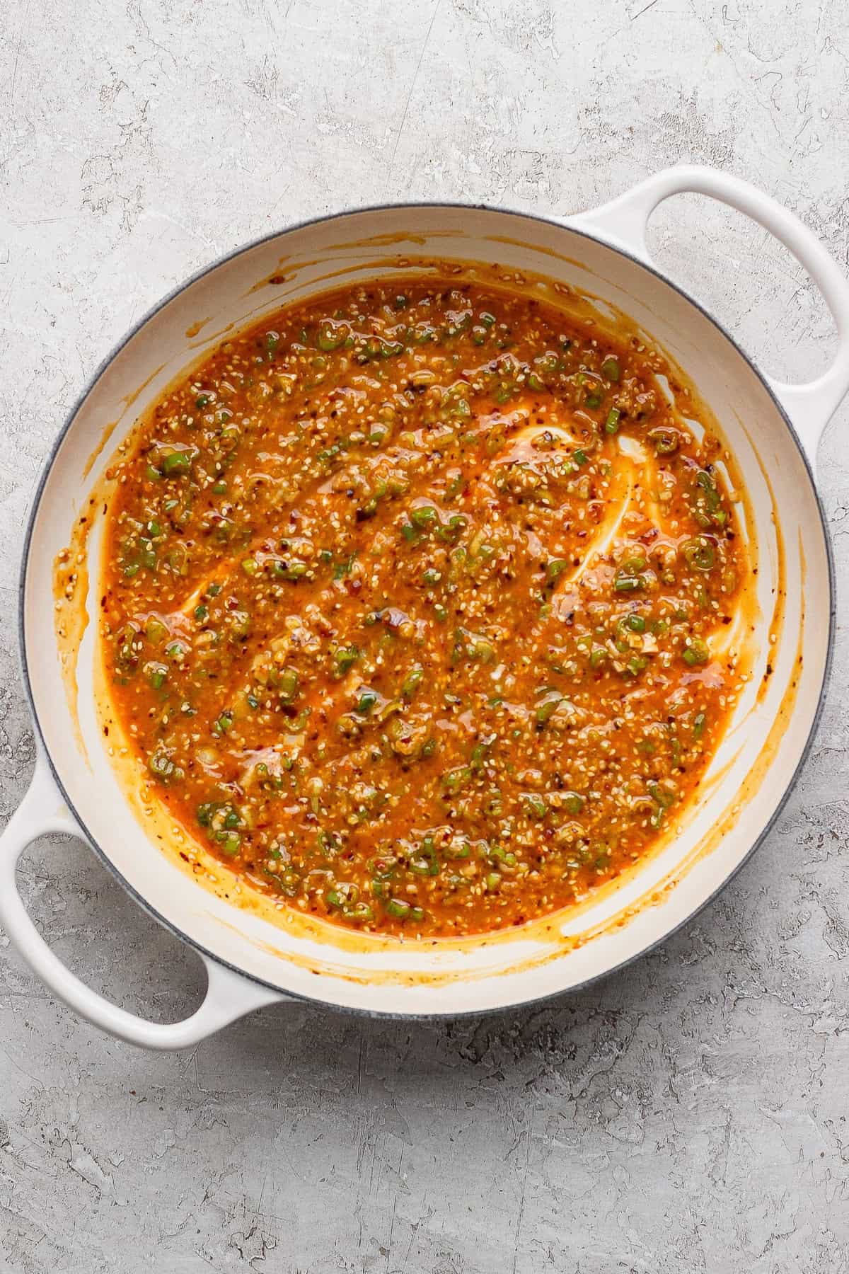 A white pot filled with a thick, orange-red sauce mixed with chopped green herbs and visible seeds, sitting on a textured light surface.