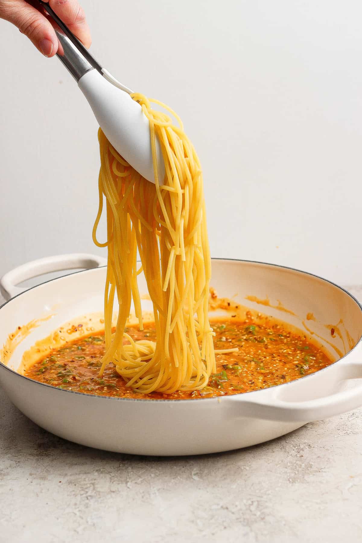A hand uses tongs to lift cooked spaghetti over a white pan filled with tomato-based sauce on a light surface.