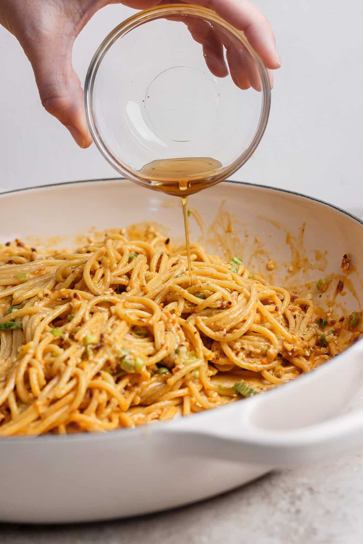A hand pours liquid from a small glass bowl onto cooked spaghetti in a white pan.